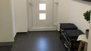 Un couloir avec une porte blanche ornée de quatre fenêtres carrées, un banc noir, une petite table avec une plante et un porte-chaussures.