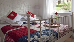 Un lit bien fait, recouvert d’une couette rouge et blanche, avec une tête de lit blanche, et une fenêtre donnant sur une maison et un jardin.