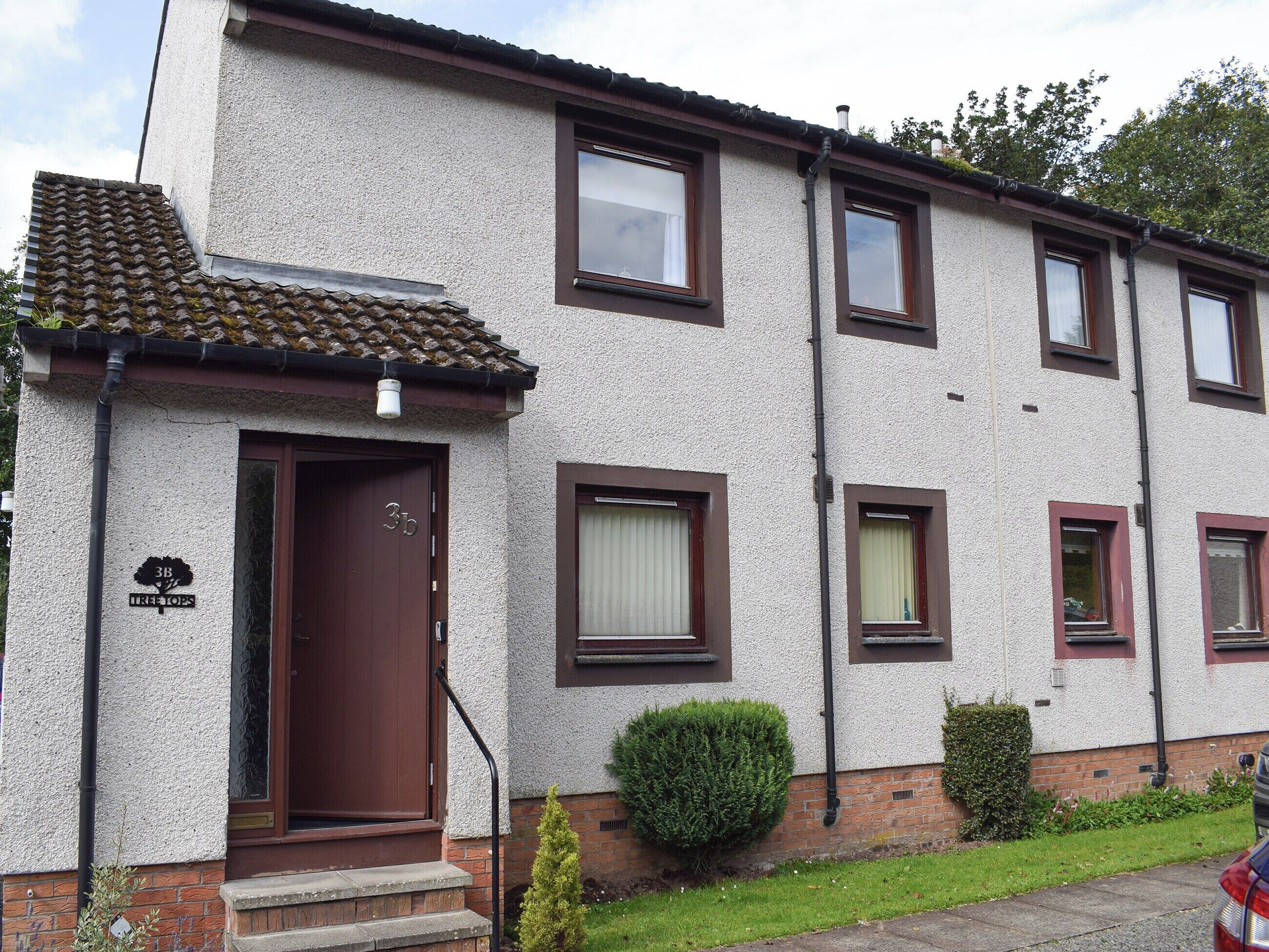 A two-story residential building with a tiled roof, a brown door, and a small front garden.