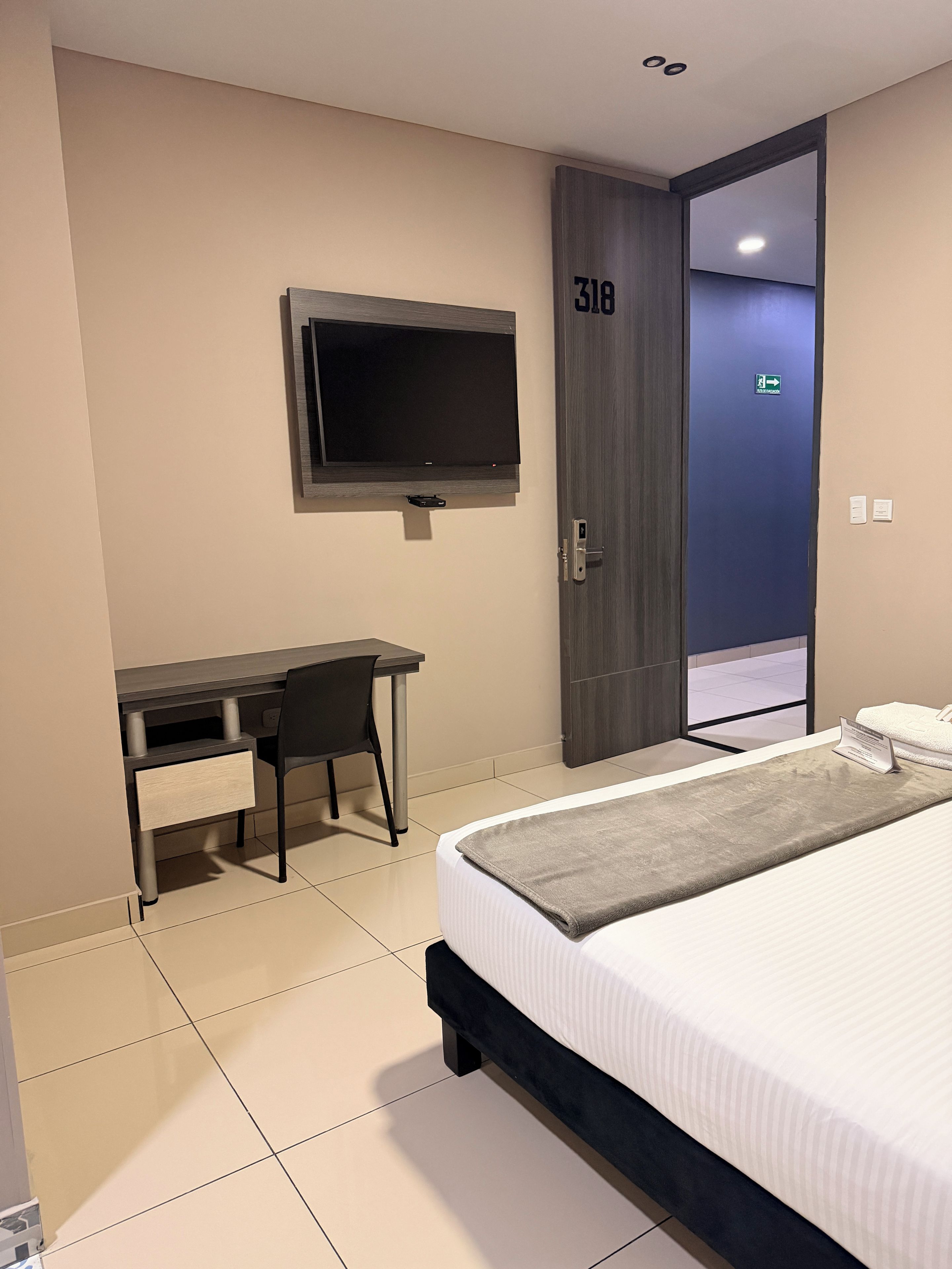 A hotel room with a bed, a desk with a chair, a television mounted on the wall, and a door with a number.