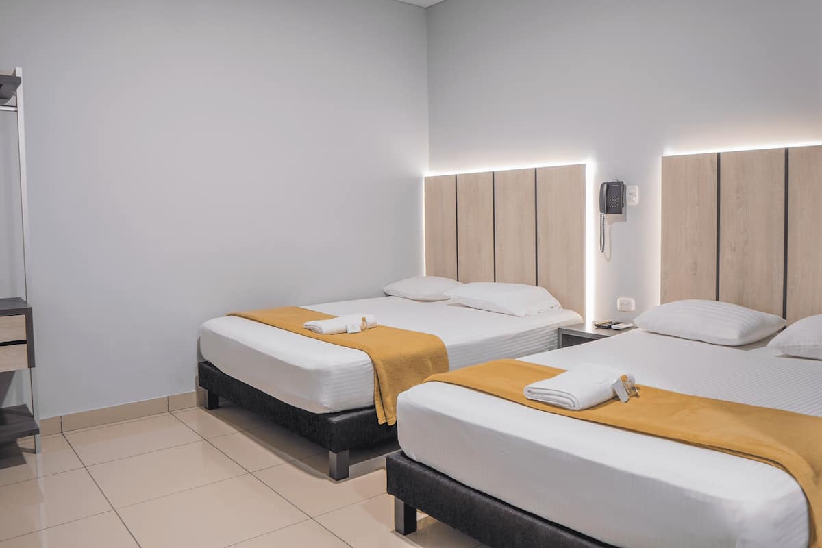 A hotel room with two beds, each with a wooden headboard, white bedding, and a mustard-colored throw blanket. There is a bedside table with a phone and a lamp on one side.