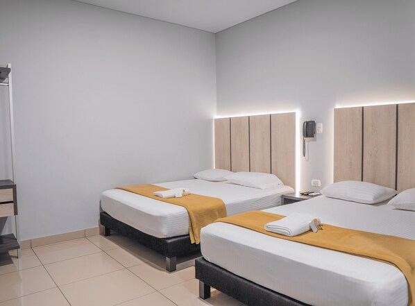 A hotel room with two beds, each with a wooden headboard, white bedding, and a mustard-colored throw blanket. There is a bedside table with a phone and a lamp on one side. - Hotel Nabú (Valledupar)