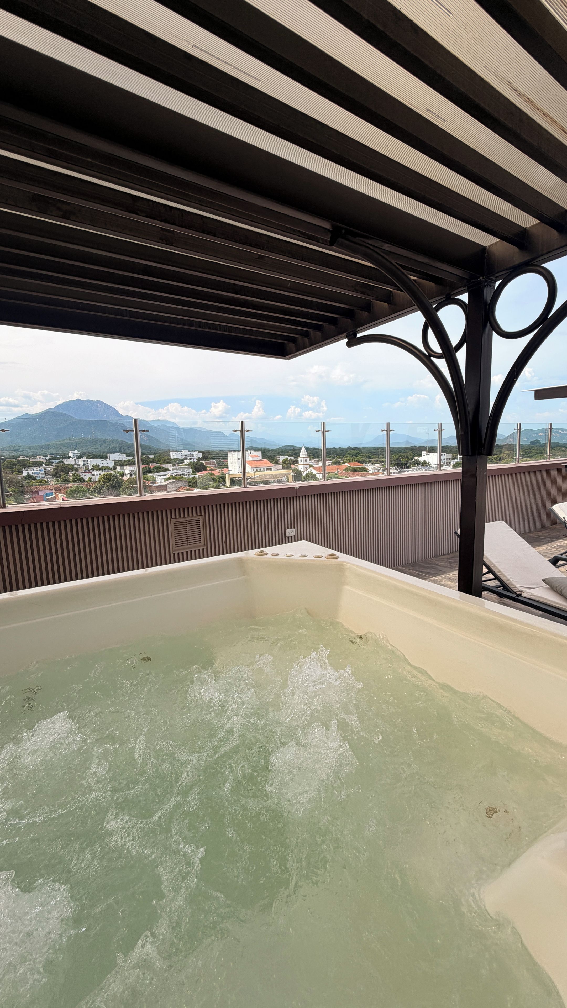A rooftop terrace with a hot tub, a view of a cityscape, and a mountainous background.