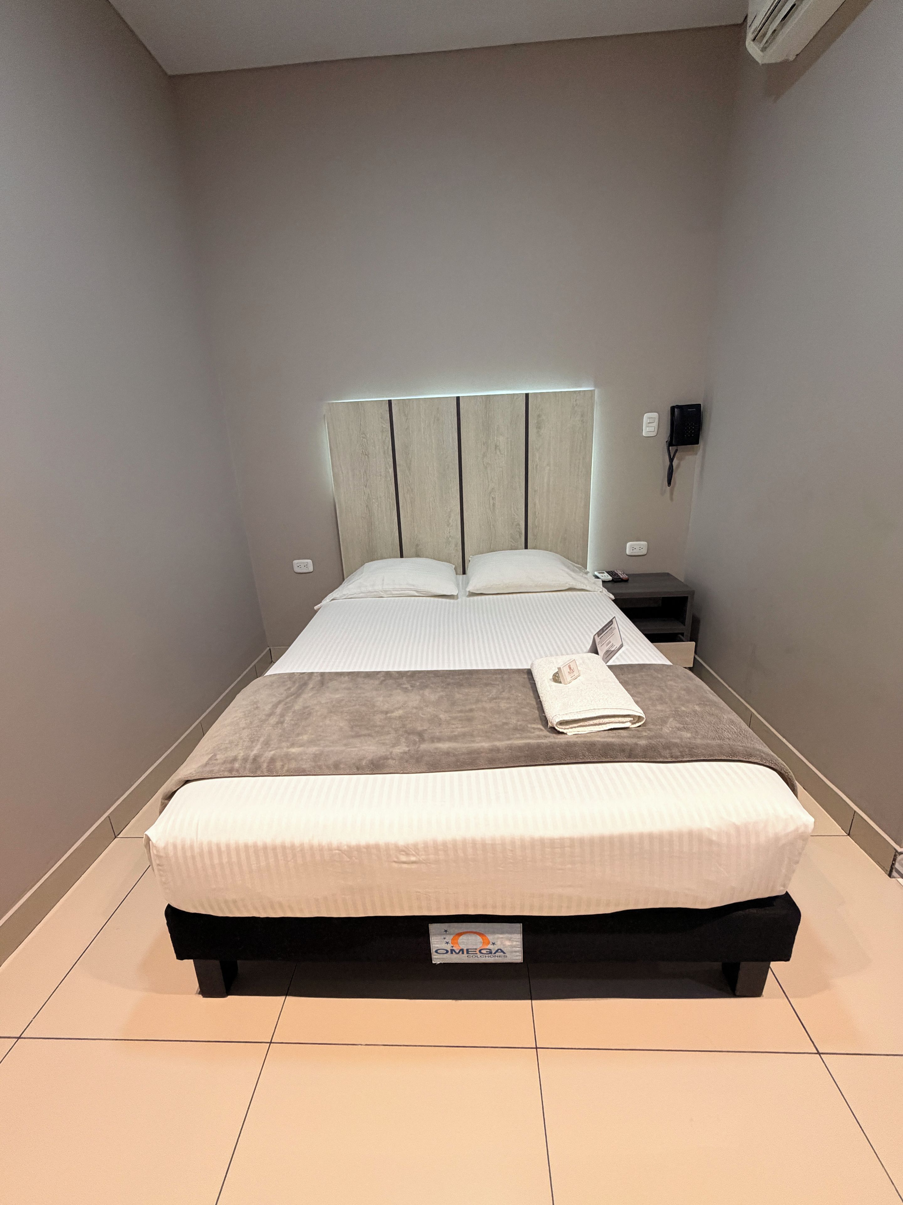 A neatly made bed with a headboard, a bedside table with a lamp, and a wall-mounted phone.