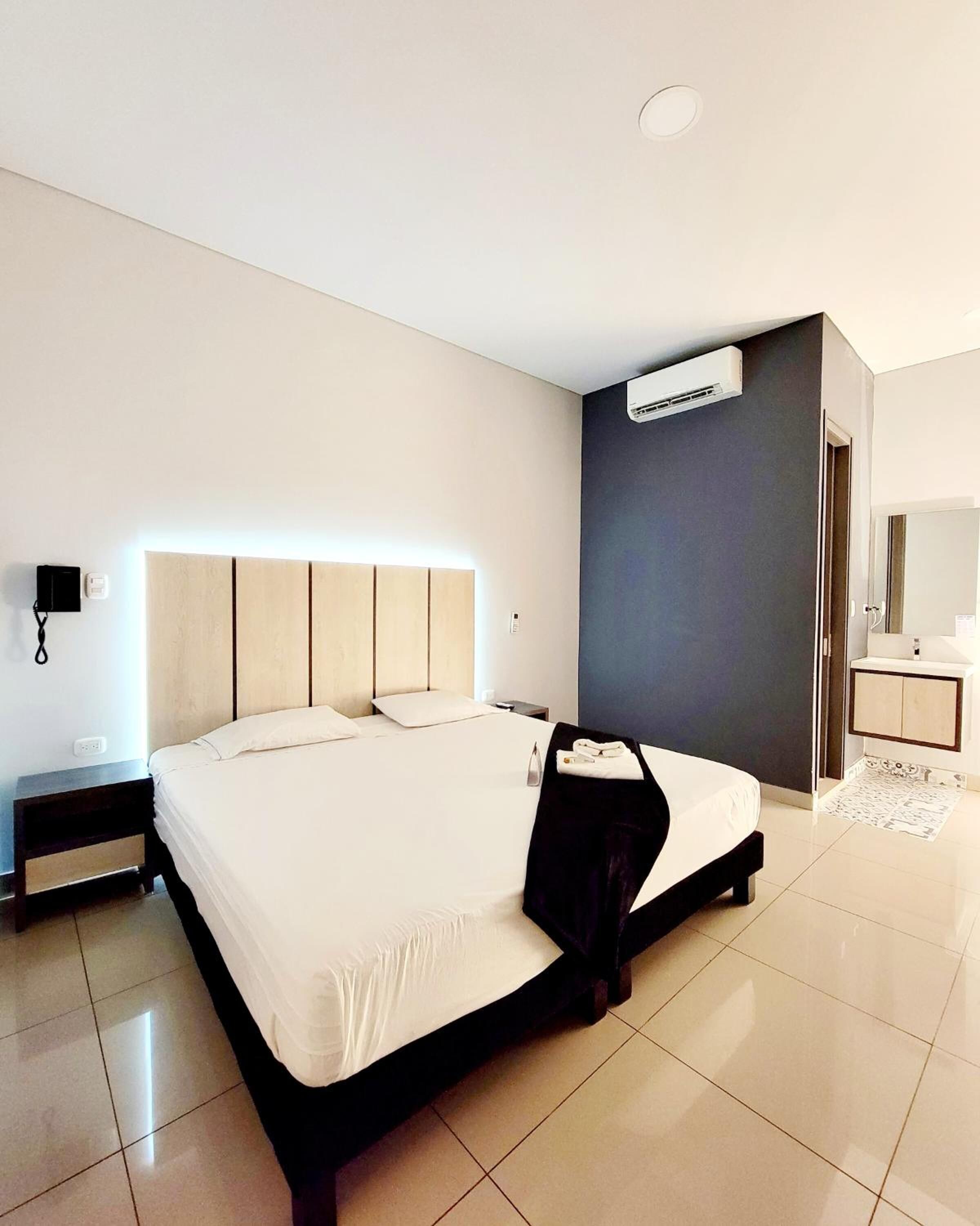 A modern hotel room with a large bed, bedside tables, a bedside lamp, an air conditioning unit, and a bathroom with a mirror and sink.