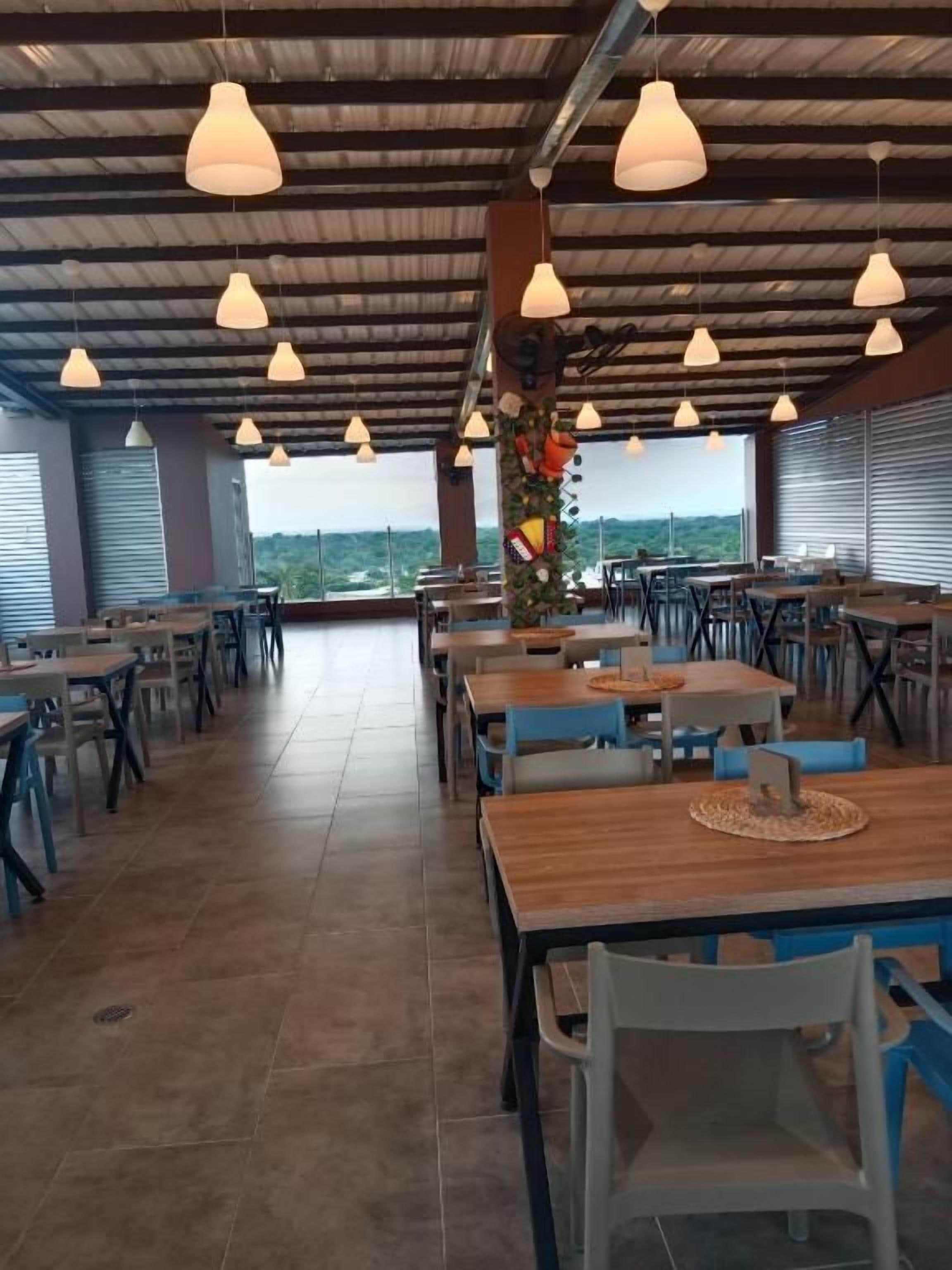 Un comedor interior con varias mesas y sillas, techo alto con luces colgantes y vistas a través de amplios ventanales.