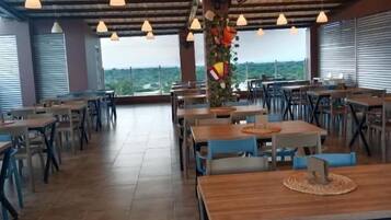 An indoor dining area with multiple tables and chairs, a high ceiling with hanging lights, and a view through large windows.