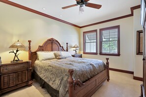 A bedroom with a large bed, two bedside tables, a ceiling fan, and two windows with blinds.