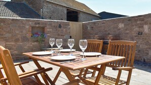Una mesa de madera al aire libre con platos, copas de vino y cubiertos, rodeada de sillas de madera, frente a una pared de piedra y un edificio rústico al fondo.