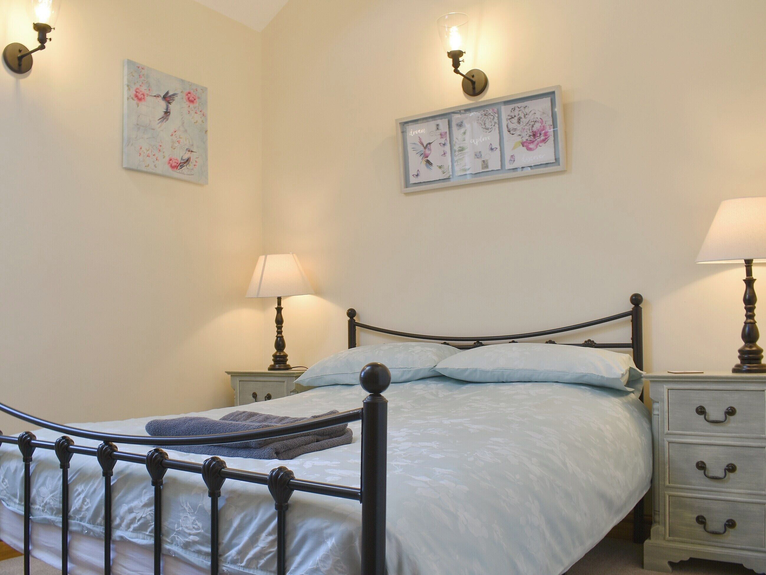 Una cama bien tendida con edredón blanco y dos almohadas, flanqueada por mesitas de noche con lámparas y cuadros en la pared.