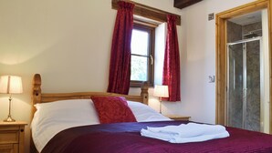 Una cama bien hecha con edredón rojo y blanco, dos lámparas de noche, una ventana con cortinas rojas y una puerta de madera que da a una zona de ducha.