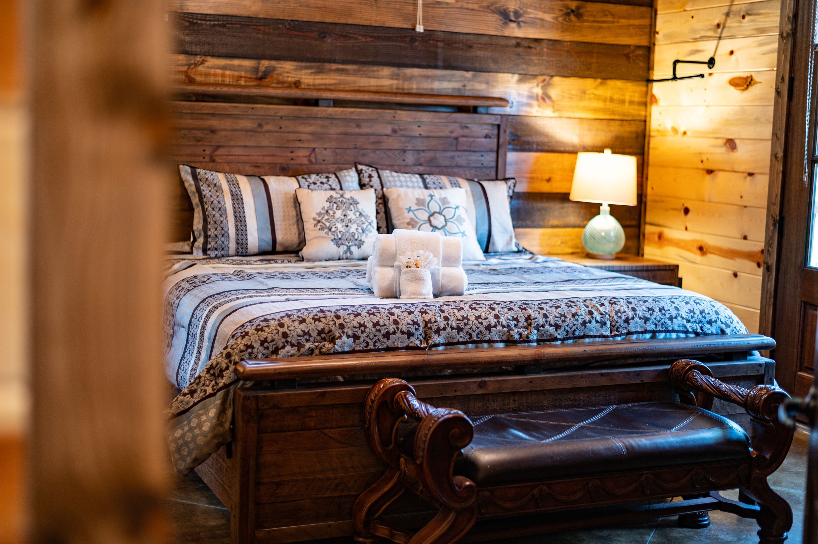 A wooden-framed bed with patterned bedding and decorative pillows, a bedside table with a lamp, and a wooden bench at the foot of the bed.