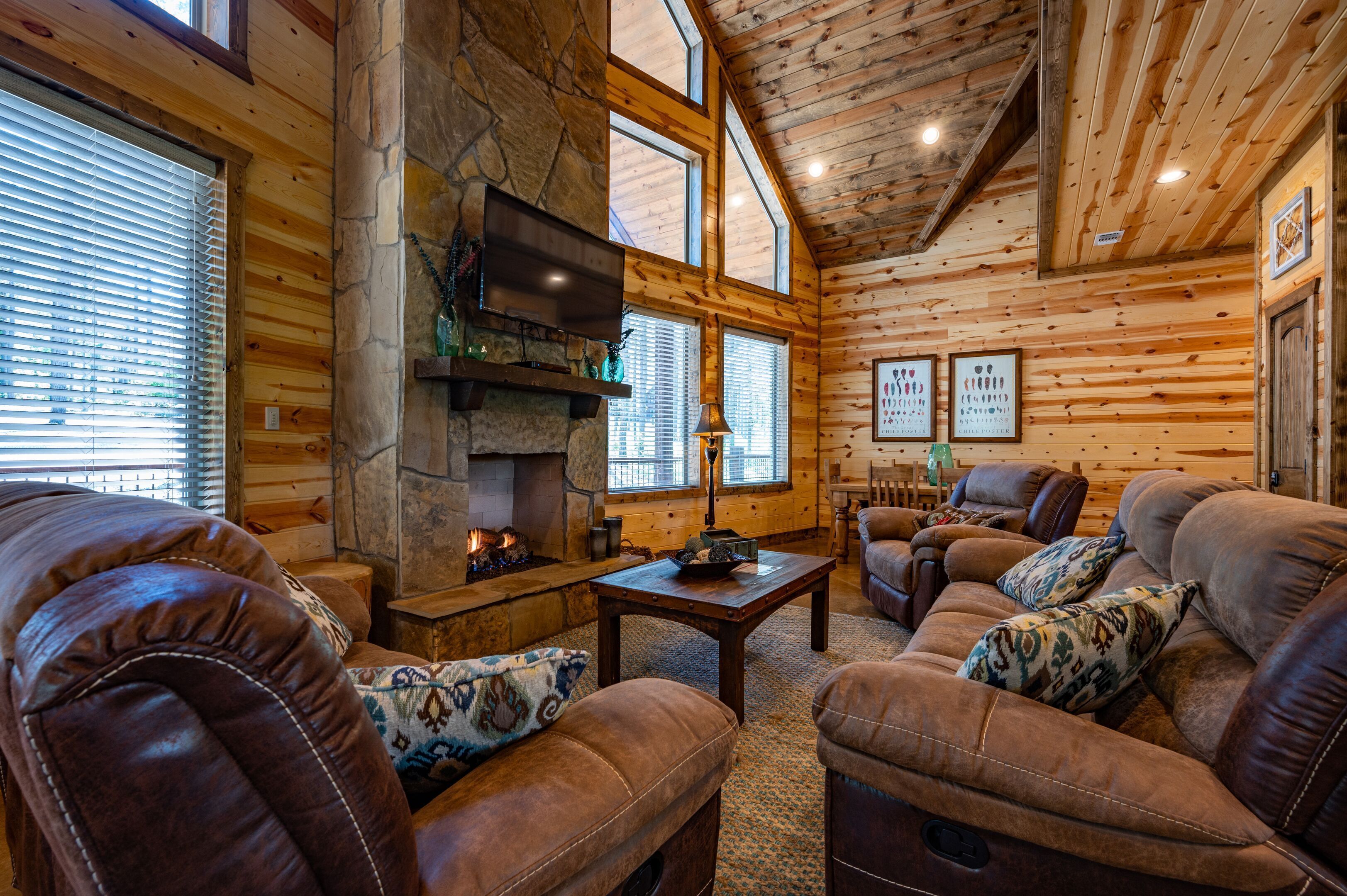 A cozy log cabin interior with leather sofas, a stone fireplace, wooden walls, and large windows.