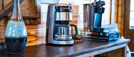 Una cafetera sobre una encimera de madera, con un jarrón y una tetera al fondo.