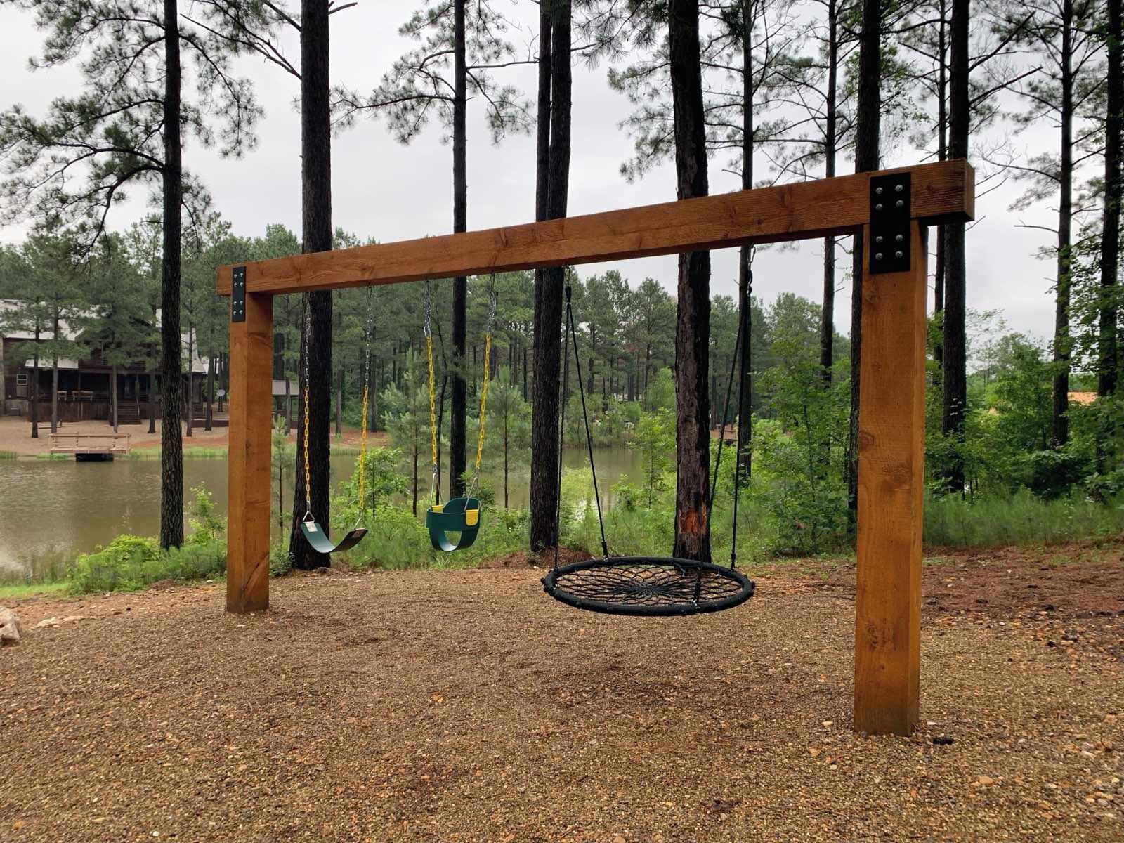A wooden swing set with two swings, a metal ring swing, and a gravel ground.