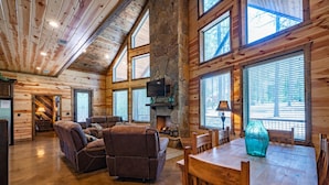 A log cabin interior with a stone fireplace, wooden furniture, and large windows.