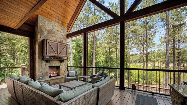 A covered deck with wicker furniture, a stone fireplace, and a view of a forest.