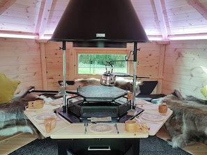 A rustic wooden interior with a wood stove, a dining table set with plates and cutlery, and a window with a view of greenery.