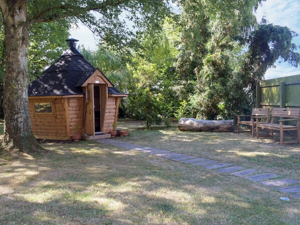 A wooden cabin with a door open, surrounded by trees and a stone pathway.