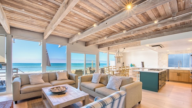 A spacious living area with a wooden ceiling, large windows overlooking the ocean, a kitchen with a central island, and a seating area with sofas and a striped chair. - 35679 Beach Rd ocean front newly remodeled 2025
 Dana Point Permit STR14-0101 (Dana Point)