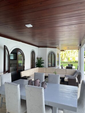 A covered outdoor seating area with a sofa, dining table, and chairs, featuring arched windows and a view of the garden. - Villa for Vacation Sosua next to the Beach (Sosua)