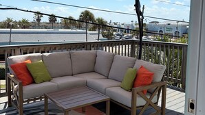 A covered outdoor seating area with a cushioned sofa and a table, a patterned rug, and a view of a parking lot and buildings in the background. -  Sleeps 12•Huge PrivateDeck•Beach Gear•Pool Table•Steps to Ocean•Trailer parking (Ormond Beach)