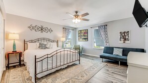 A bedroom with a bed, a ceiling fan, a TV, a sofa, and a window with curtains. -  Sleeps 12•Huge PrivateDeck•Beach Gear•Pool Table•Steps to Ocean•Trailer parking (Ormond Beach)
