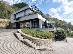 Un edificio moderno con una escalera de piedra que conduce a una entrada de vidrio, rodeado de follaje y una zona pavimentada.
