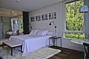 A modern bedroom with a large bed, a small table, a bench, and a view of greenery through a window.