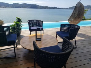 A deck with wicker chairs and a table overlooking a pool and a lake.