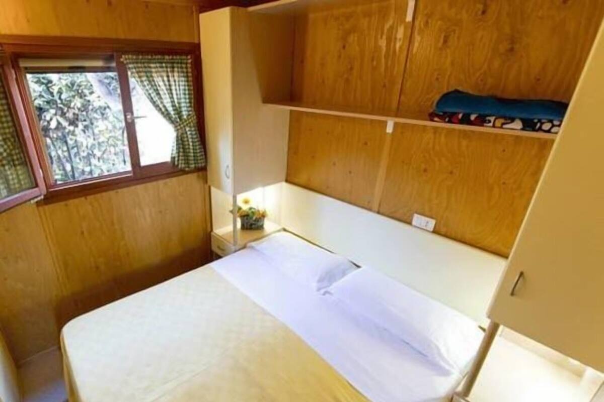 Ein kleines, gemütliches Zimmer mit einem Einzelbett, Holzwänden, einem Fenster mit Vorhängen und einem kleinen Tisch mit einer Lampe.