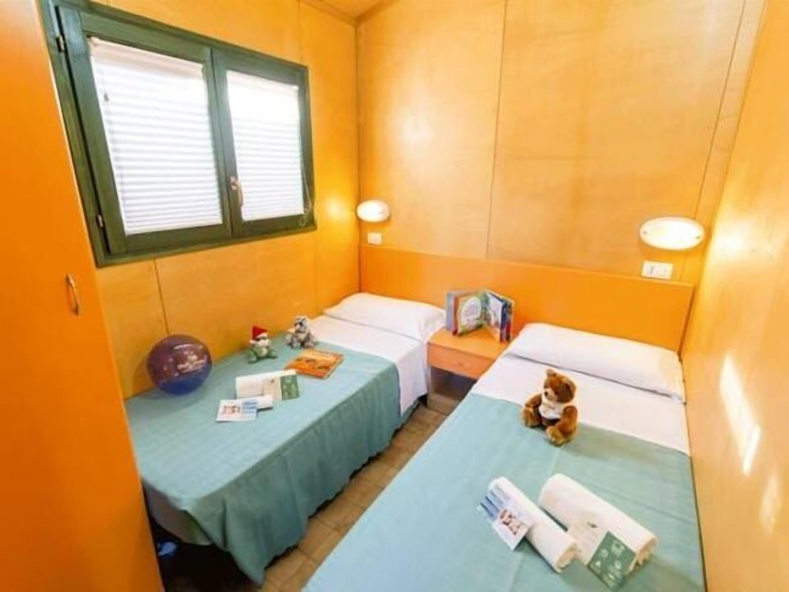A small, single-room accommodation with two beds, a window with blinds, a bedside table with a lamp, and a small shelf with items.