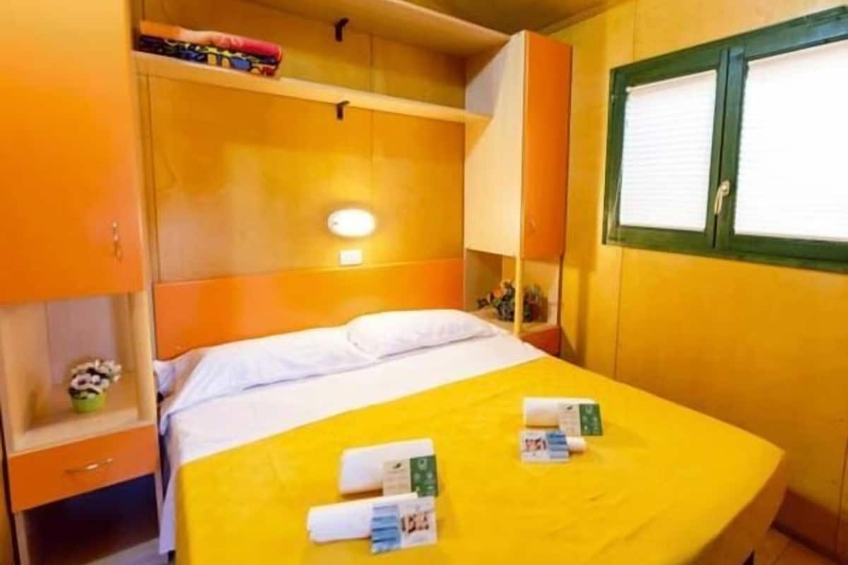 Ein Einzelbett mit gelber Bettdecke, ein Nachttisch mit Lampe und ein Fenster mit Jalousien.