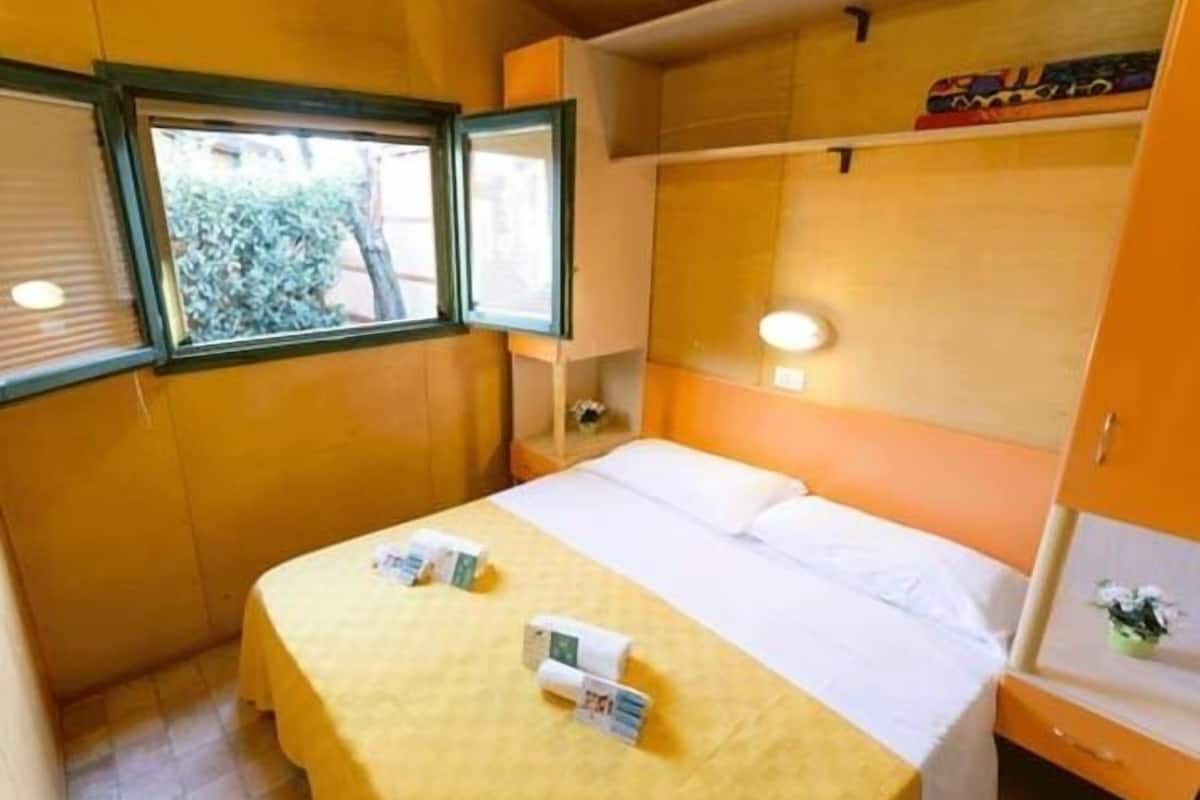 Ein kleines, gemütliches Zimmer mit einem Einzelbett, einem Fenster mit Jalousien, einem Nachttisch mit Lampe und einem Regal über dem Bett.