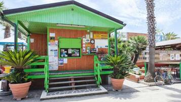 A small, green-roofed building with a wooden facade, steps leading up to a green door, and potted plants around.