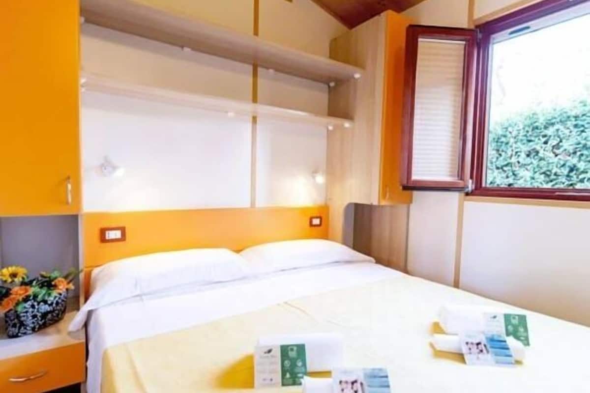 Ein Hotelzimmer mit Bett, Nachttisch, Fenster mit Jalousien und Blick ins Grüne.