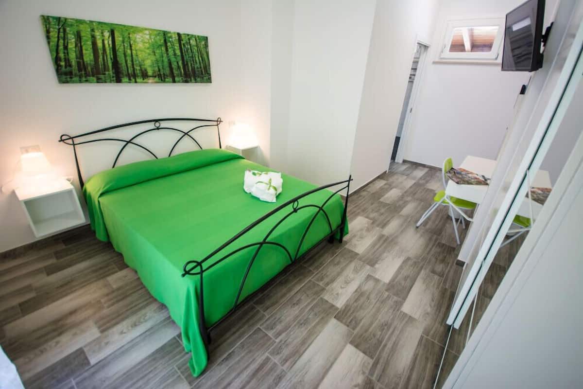 A bedroom with a bed, bedside lamps, a green bedspread, a painting of a forest, and a TV on the wall.