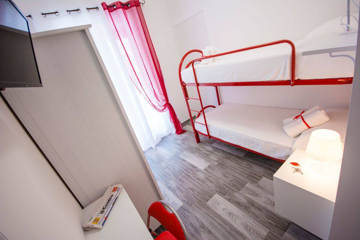 A dormitory room with a bunk bed, a small bedside table with a lamp, a chair, and a window with curtains.