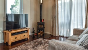 A living room with a television on a wooden stand, a fireplace with logs, a sofa, and a large window with curtains. - 2 bedroom accommodation in Yanwath, near Penrith (Yanwath, near Penrith)