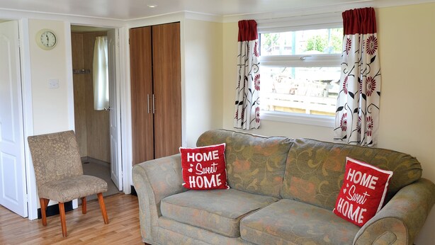 A living room with a sofa, two pillows, a chair, wooden flooring, a wardrobe, a clock, and a window with curtains. - 2 bedroom accommodation in Stalham (Stalham)