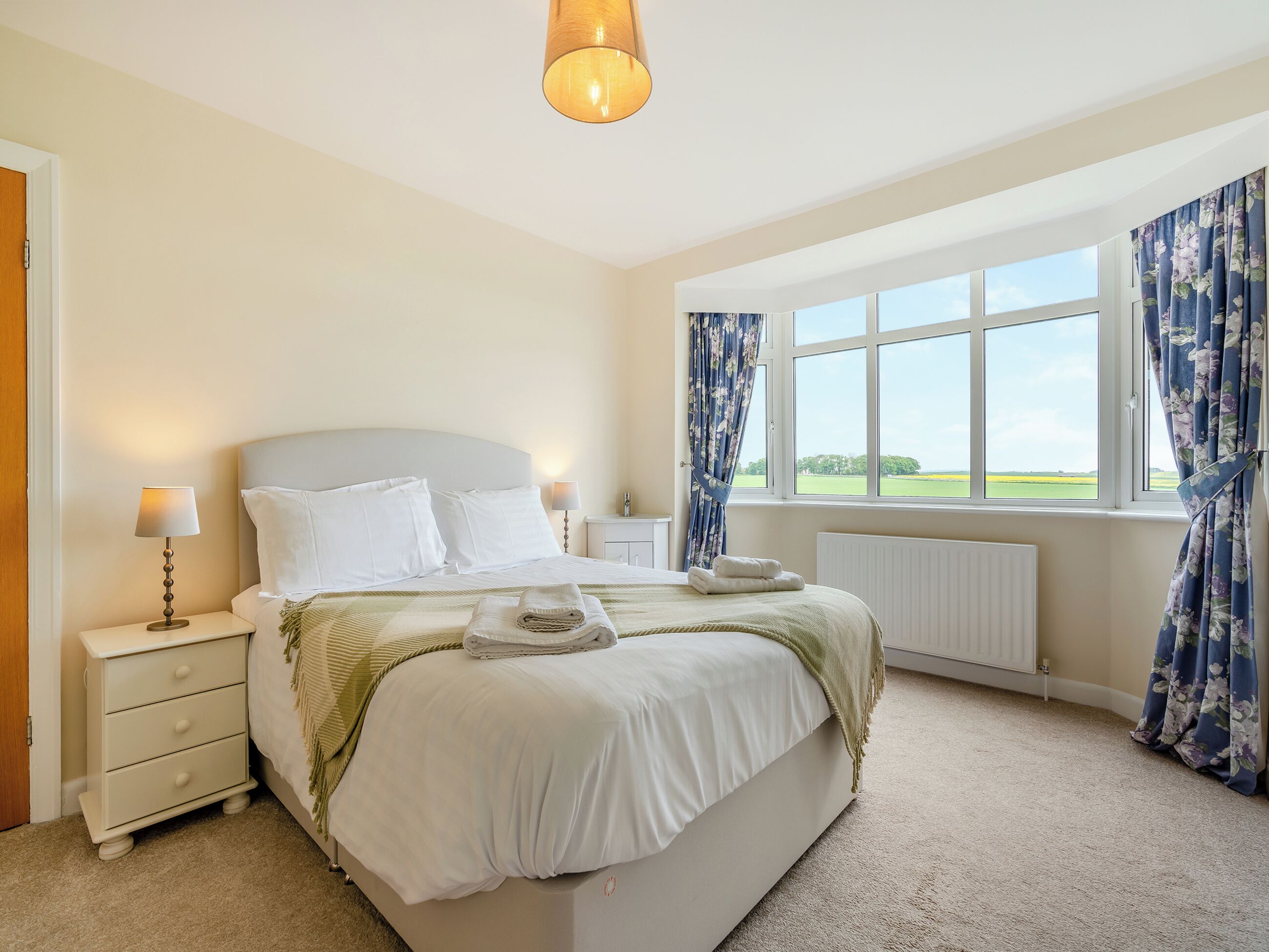 A bedroom with a large bed, two bedside tables, a window with curtains, and a view of the countryside.