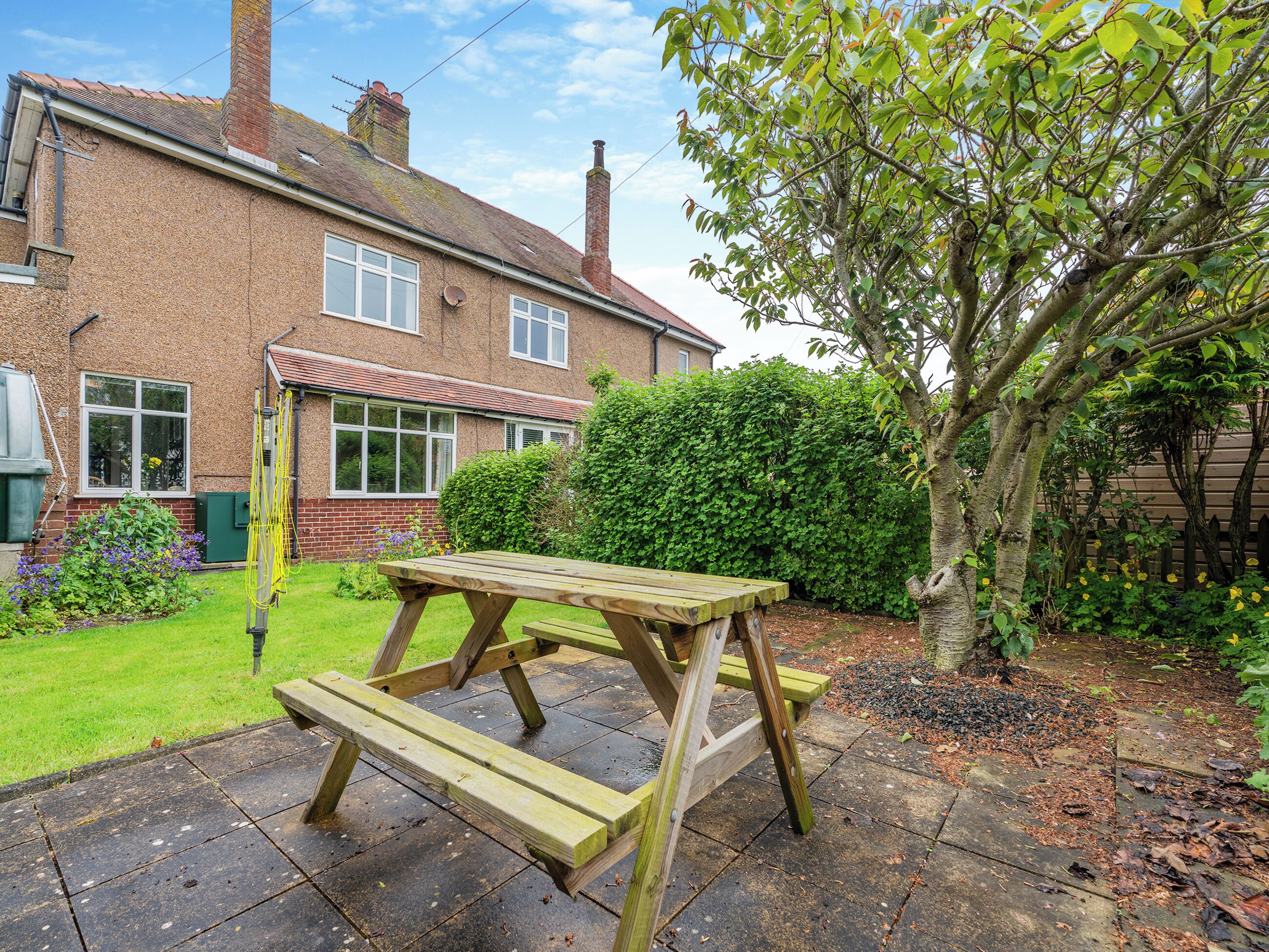 A wooden picnic table in a garden with a brick house in the background.
