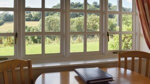 A room with a large window overlooking a green landscape, a wooden table with a book, and two wooden chairs. - 1 bedroom accommodation in Sidbury, near Sidmouth (Sidbury, near Sidmouth)