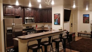 A modern kitchen with dark wooden cabinets, stainless steel appliances, a central island with bar stools, and a dining area with a sofa and a TV. - Slickrock Sands - Sleeps 8! Pet/bike friendly. (Moab)