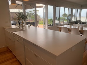 A modern kitchen with a large island, a dining area with chairs, and a view of the ocean through large windows. - Fern - Ocean Views, Middle Of Town Pet Friendly (Lorne)