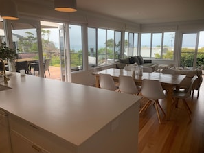 A modern kitchen with a large island, dining area with chairs and a table, and large windows offering a view of the outdoors. - Fern - Ocean Views, Middle Of Town Pet Friendly (Lorne)