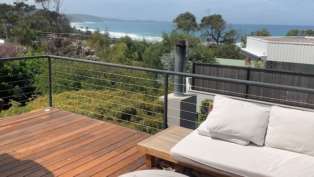 Une terrasse avec un plancher en bois, un canapé blanc avec des coussins et une table décorée de bougies. Une rambarde en métal entoure la terrasse.