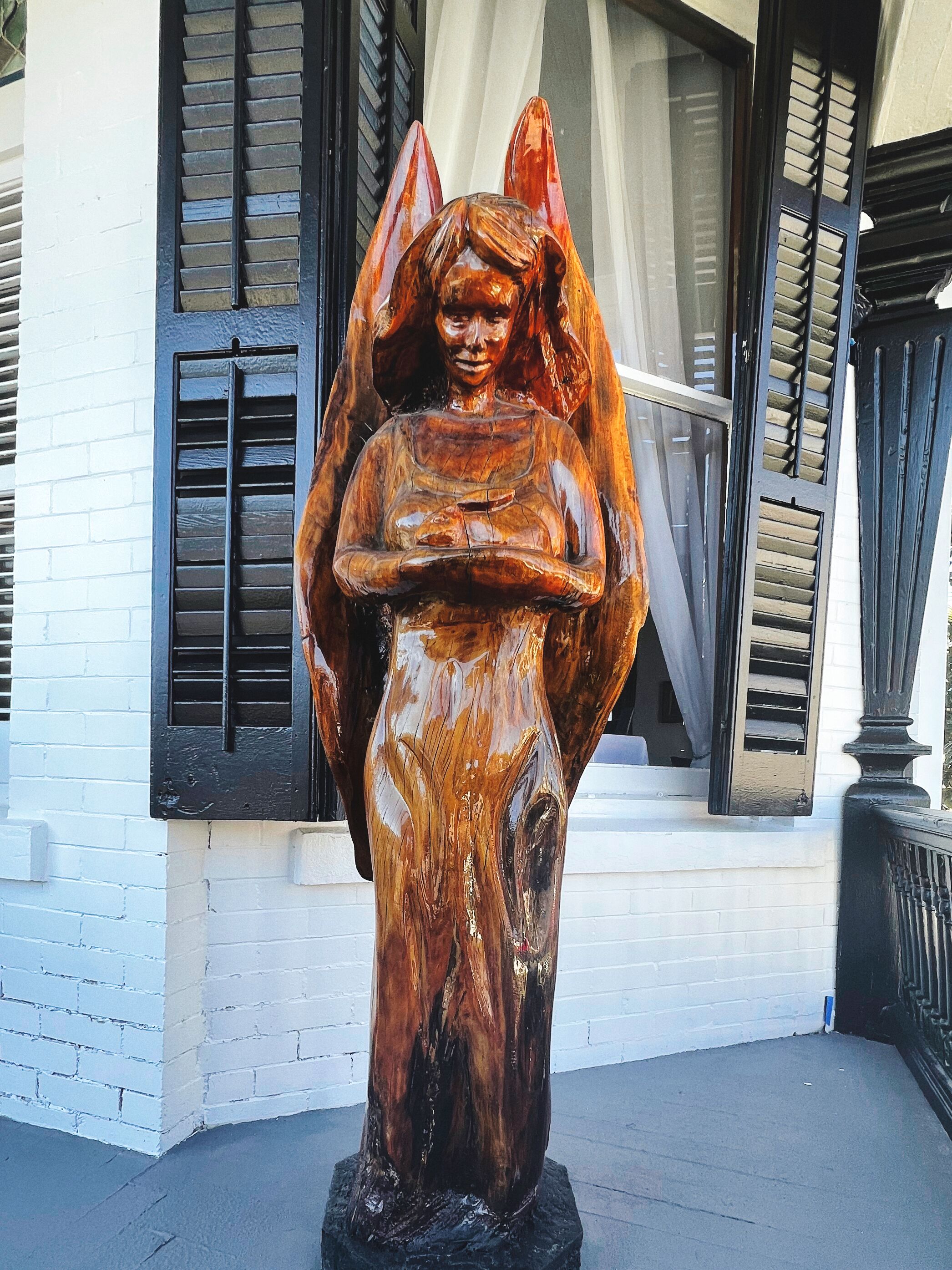 Una estatua de ángel de madera frente a un edificio con paredes de ladrillo blanco y contraventanas negras.