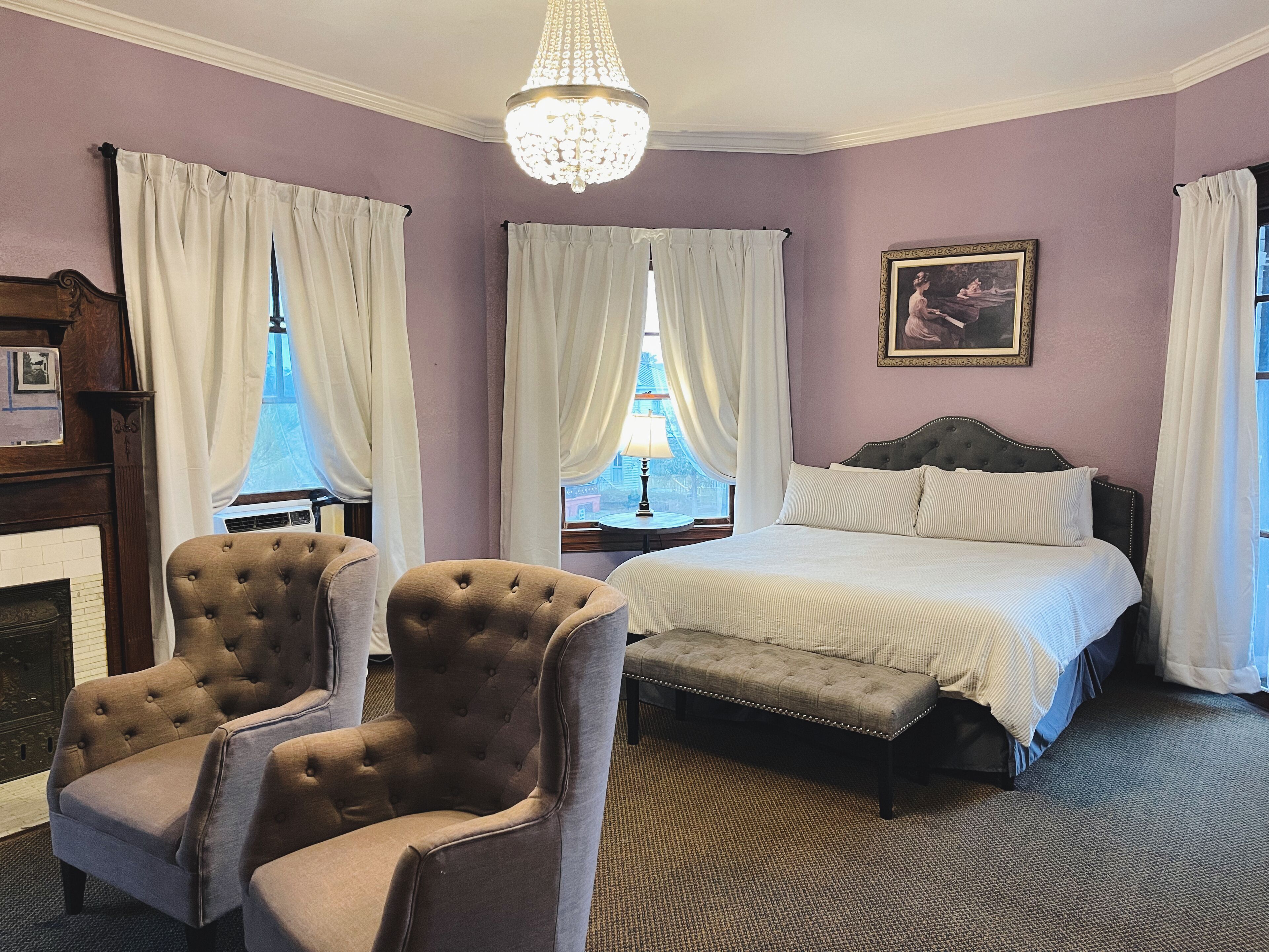 A bedroom with a bed, two armchairs, a bench, a chandelier, a fireplace, and a window with curtains.