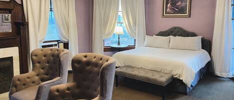 A bedroom with a bed, two armchairs, a bench, a chandelier, a fireplace, and a window with curtains.